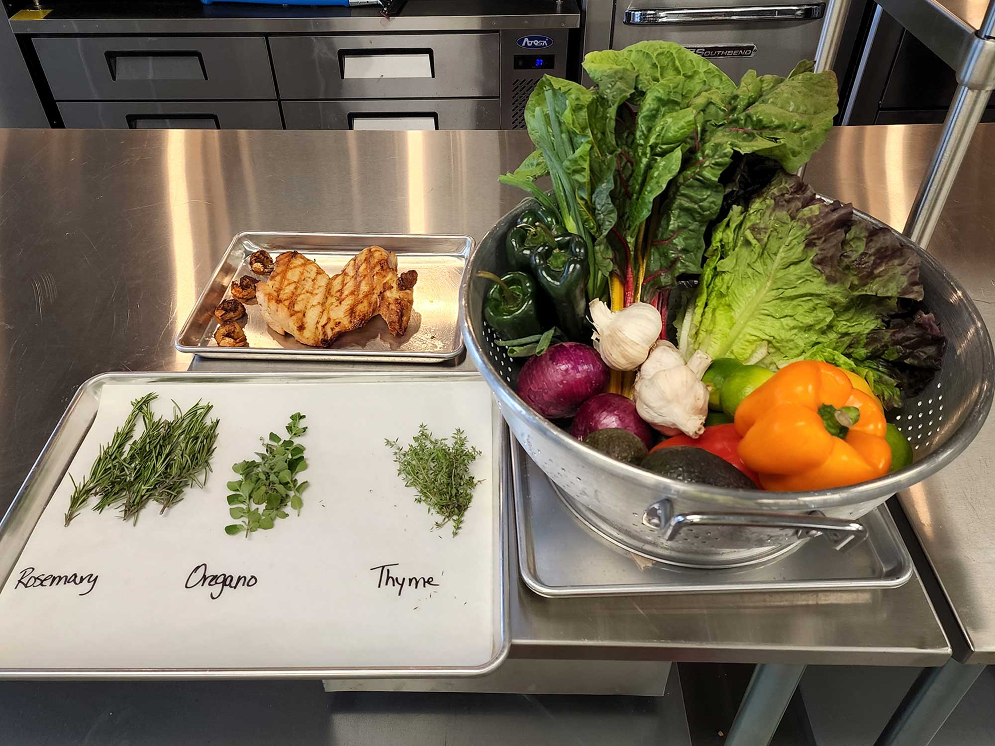 various foods are displayed on a metal industrial kitchen countertop. three bunches of herbs are labeled rosemary, oregano and thyme.