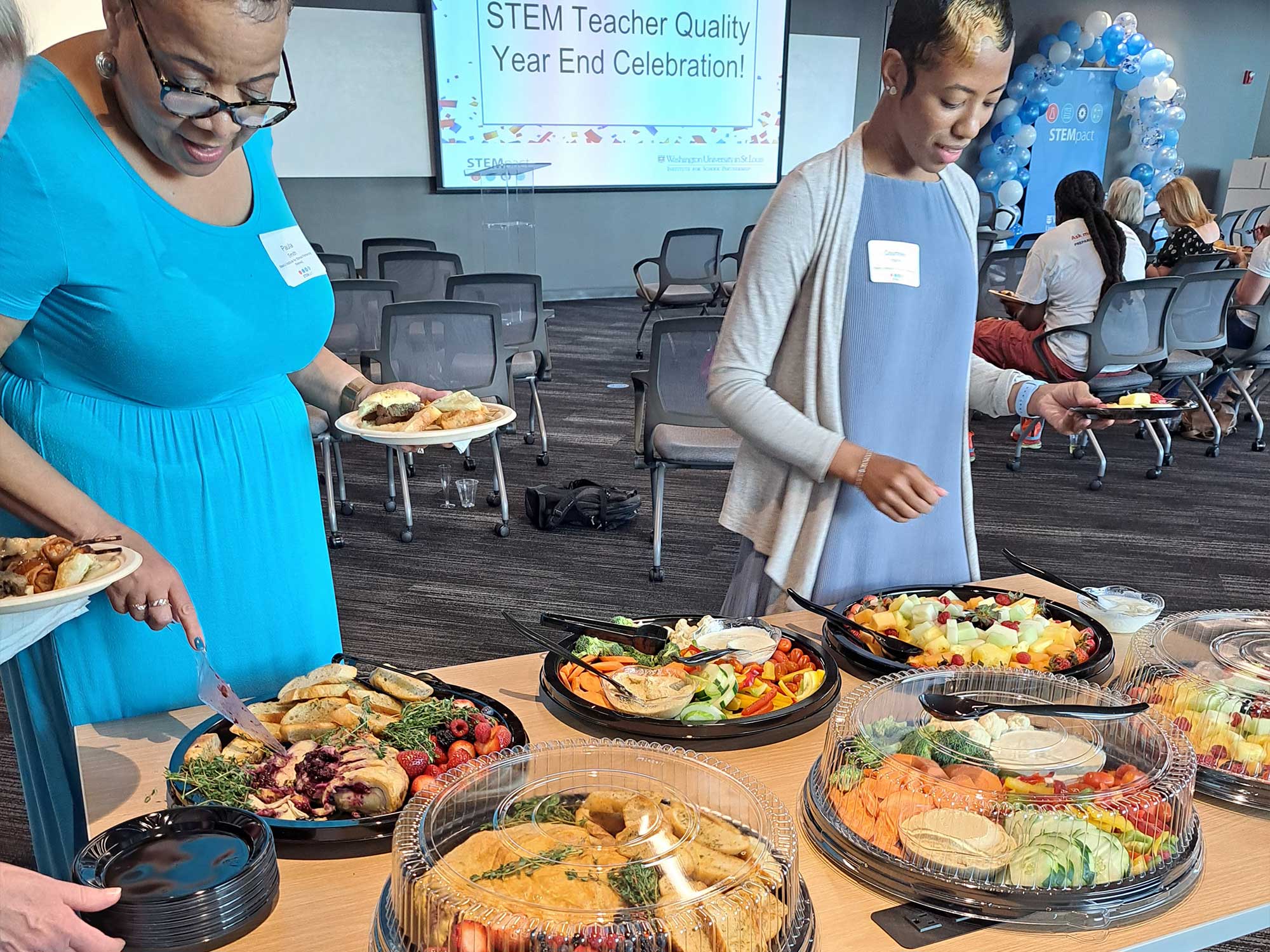 two black women serve themselves food from a table of catered platters of food. a sign in the background indicates that this is a STEM teacher appreciation event. 